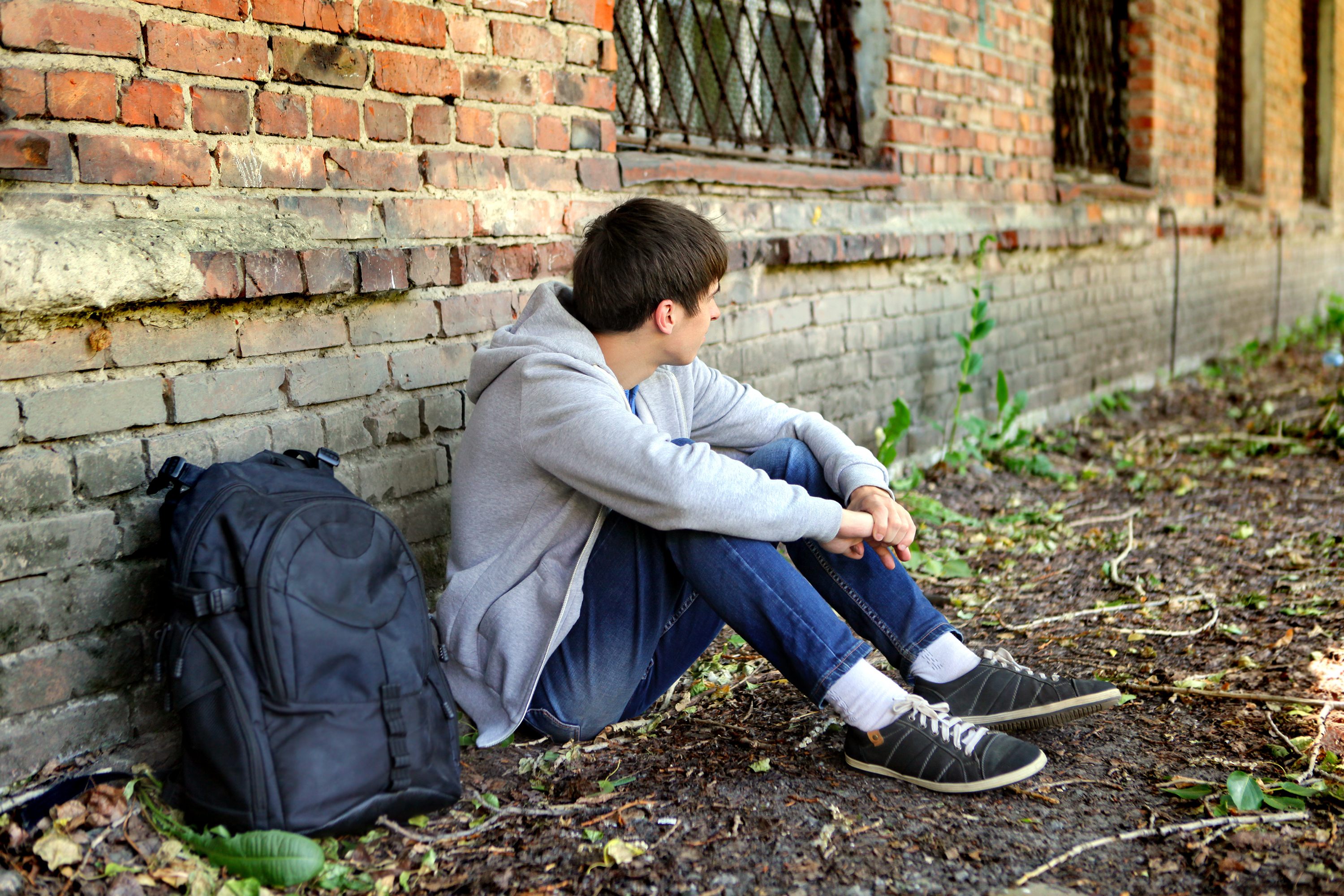 Ung mann sitter på bakken inntil murvegg, med blikket bortvendt. 